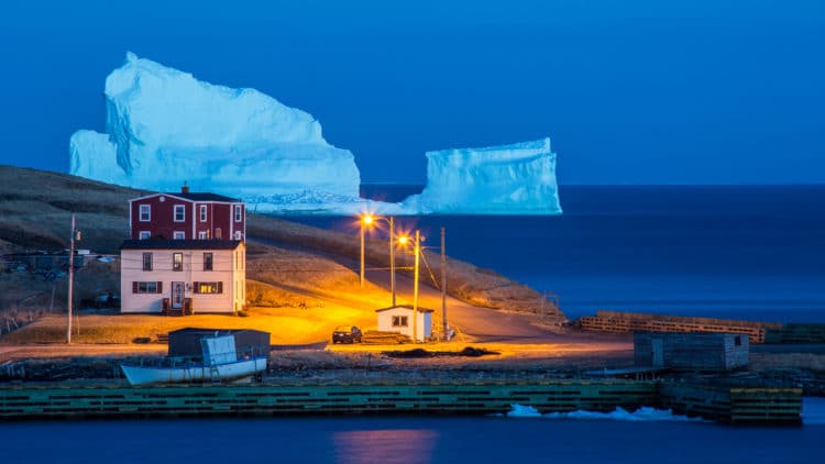A large iceberg is in the ocean near some buildings.