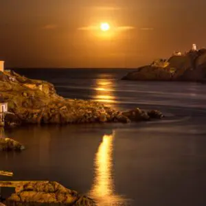 A full moon setting over the ocean with buildings in the distance.