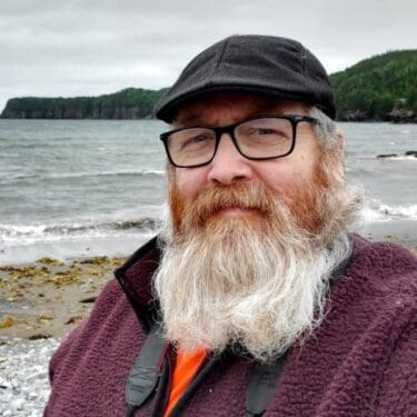 A man with long beard and glasses standing on the beach.